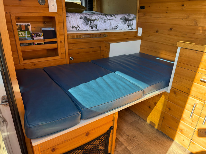 Cozy RV rental features wood walls, blue convertible seating, an overhead bunk bed, and storage for van life adventures. — photo 8