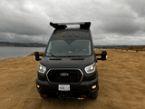 A rugged black Ford Transit RV rental, with a light bar and winch, parked on a sandy beach on a cloudy day.