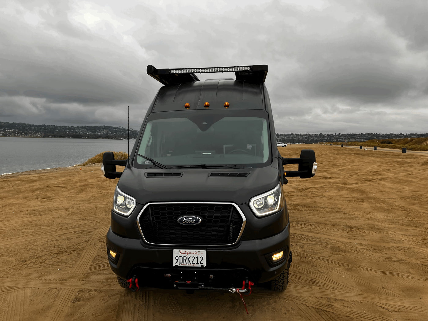 A rugged black Ford Transit RV rental, with a light bar and winch, parked on a sandy beach on a cloudy day. — photo 8