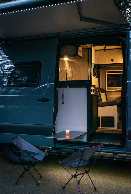 Illuminated RV rental interior visible through its open side door, showcasing a cozy setup with a candle-lit table & outdoor chairs.