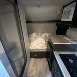 Cozy RV rental interior: featuring a compact kitchen, bed with protective wrap, and a shower/toilet bathroom on the left.