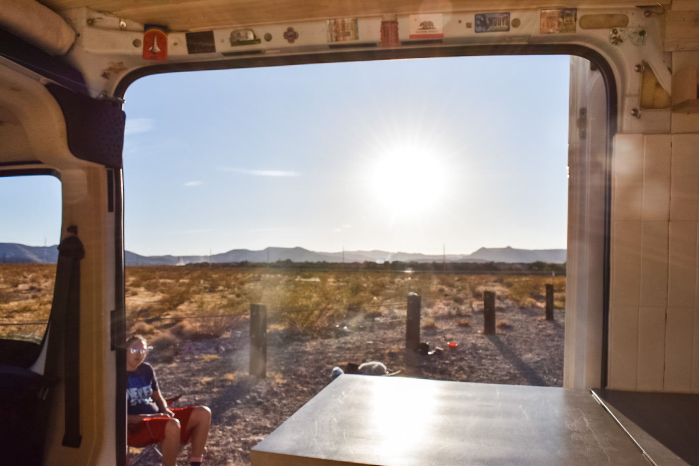 View from inside an RV rental overlooking a vast, sunny desert with mountains. A person and dog relax outside while camping.