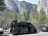 An rv rental, a Winnebago Solis camper van, parked in front of towering Yosemite Falls and granite cliffs. Perfect nature travel.