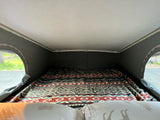 Cozy RV rental pop-top bed with vibrant patterned blanket. Windows show lush greenery outside.