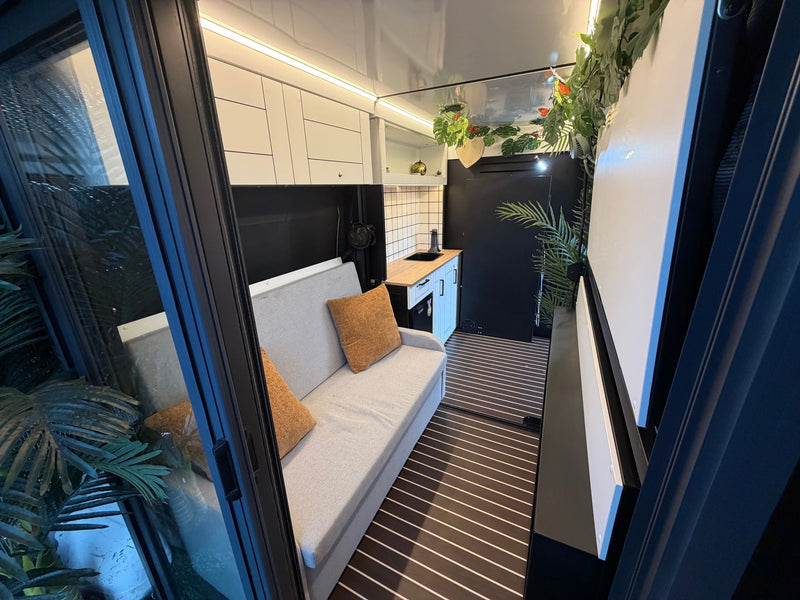 Modern rv rental interior with a cozy light grey sofa, compact kitchenette, and decorative greenery. Striped floor. — photo 18