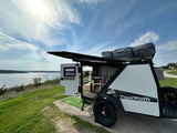 A TigerMoth camper trailer, an ideal RV rental, by a shining lake under sunny skies, with an open side for outdoor living.