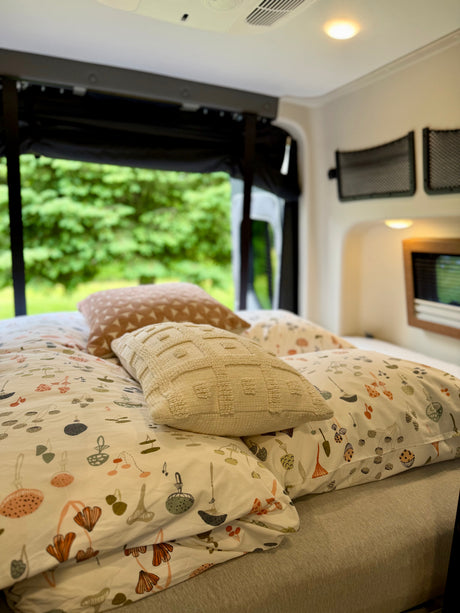 A cozy rv rental bedroom with mushroom-patterned bedding & textured pillows. Lush green trees are visible outside the window.