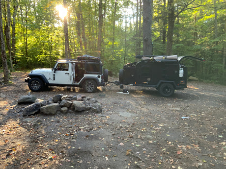 A white Jeep and black overland trailer parked at a sunny campsite with a rock fire pit. Perfect for an RV rental outdoor adventure.