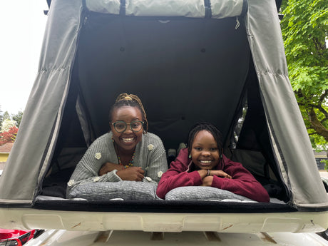 Two joyful people, an adult and child, inside an RV rental rooftop tent, smiling at the camera. Ideal for family camping trips.
