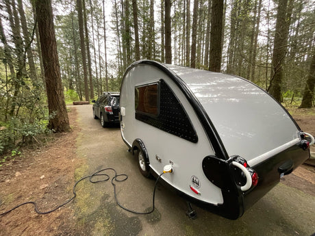 Teardrop RV rental parked in a peaceful forested campground, connected to power, with tow vehicle visible.