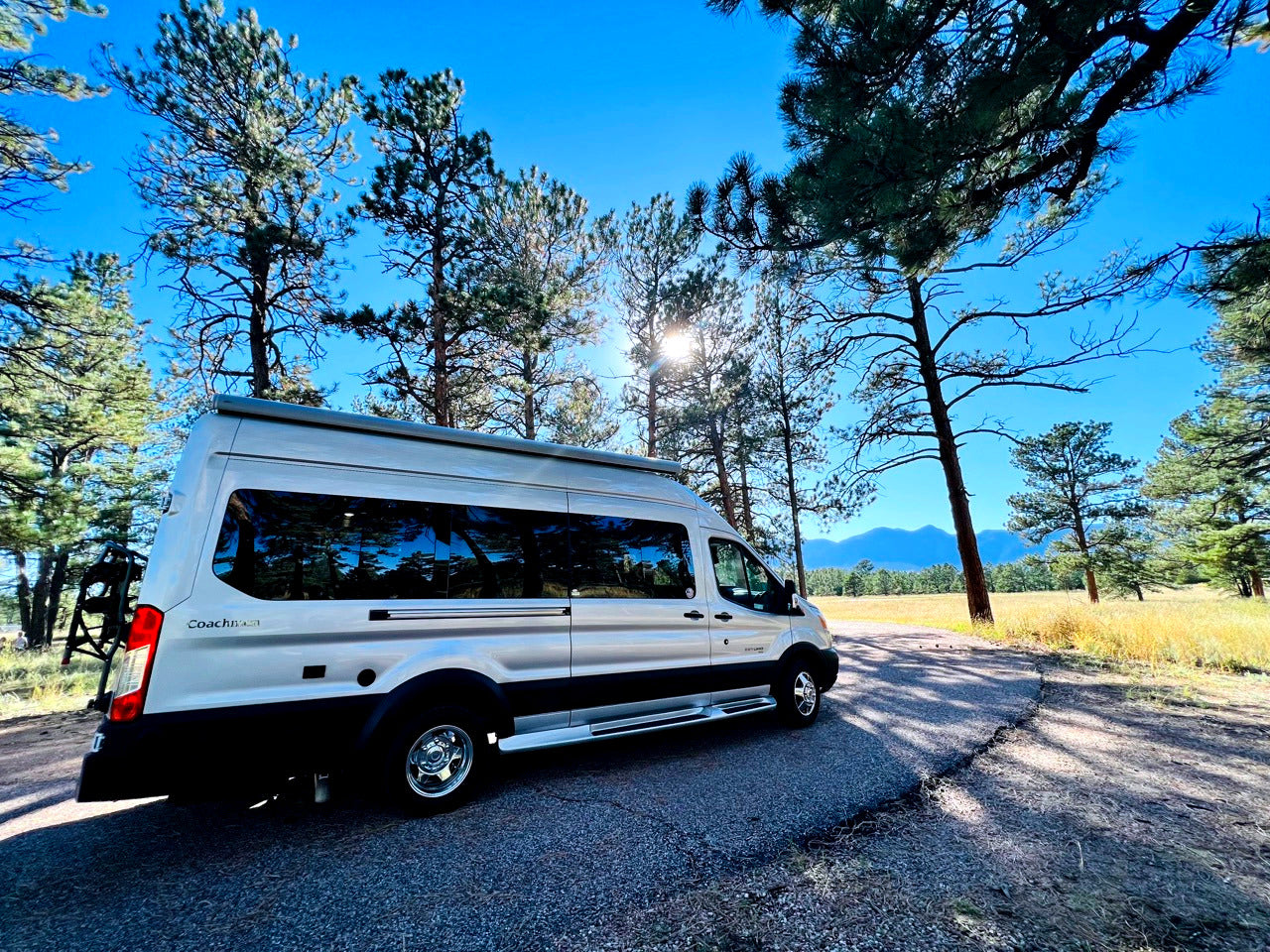 Silver RV rental camper van parked on a sunny road, surrounded by tall pine trees and distant mountains. Ideal for your next road trip.