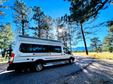 Silver RV rental camper van parked on a sunny road, surrounded by tall pine trees and distant mountains. Ideal for your next road trip.