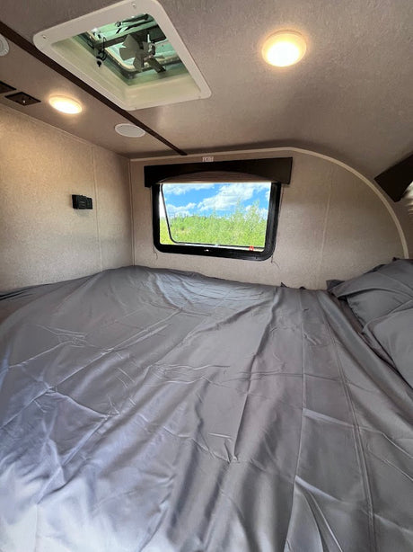 Cozy RV rental bedroom featuring a grey bed, window view of trees, ceiling vent, and bright lights.