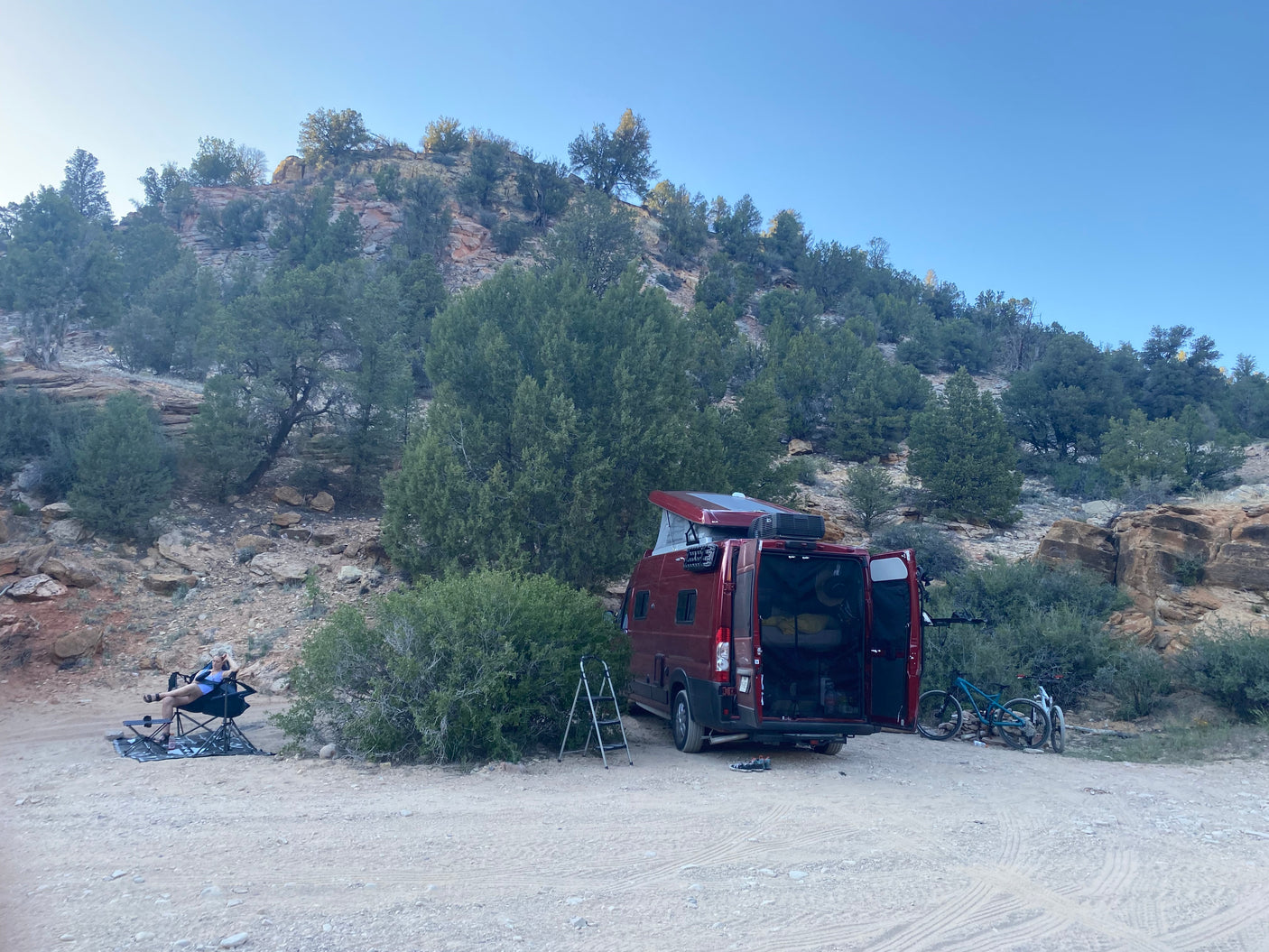 Relaxing by a red RV rental van with bikes & pop-top, parked on a dirt road by a rocky hillside.