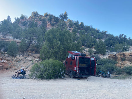 Relaxing by a red RV rental van with bikes & pop-top, parked on a dirt road by a rocky hillside.