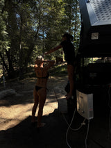 RV rental outdoor shower fun: A woman in a bikini rinses off while another assists in a sunny forest.