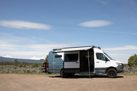 Muse & Co. rv rental camper van with awning extended, parked on a dirt road with snowy mountains and trees in the background. Ready for adventure!