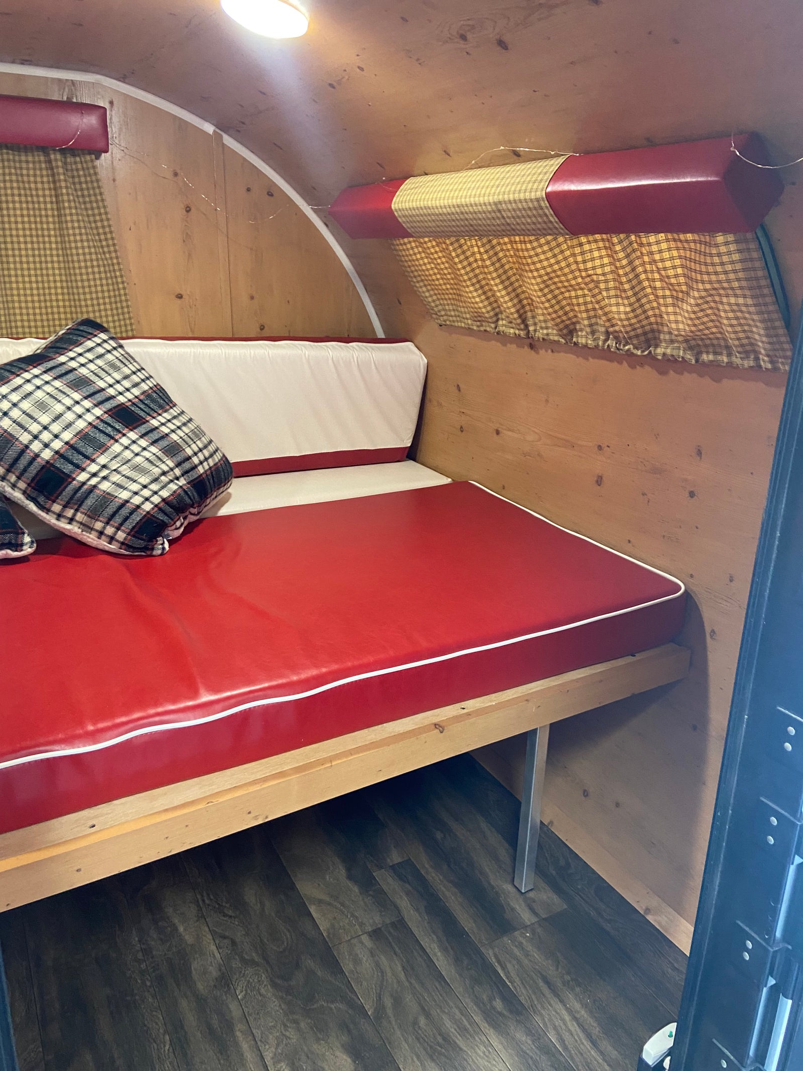 Cozy RV rental interior with a red and white upholstered bench bed, plaid pillow, wooden walls, and gingham curtains.