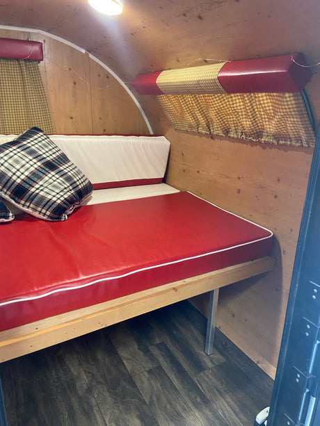 Cozy RV rental interior with a red and white upholstered bench bed, plaid pillow, wooden walls, and gingham curtains.