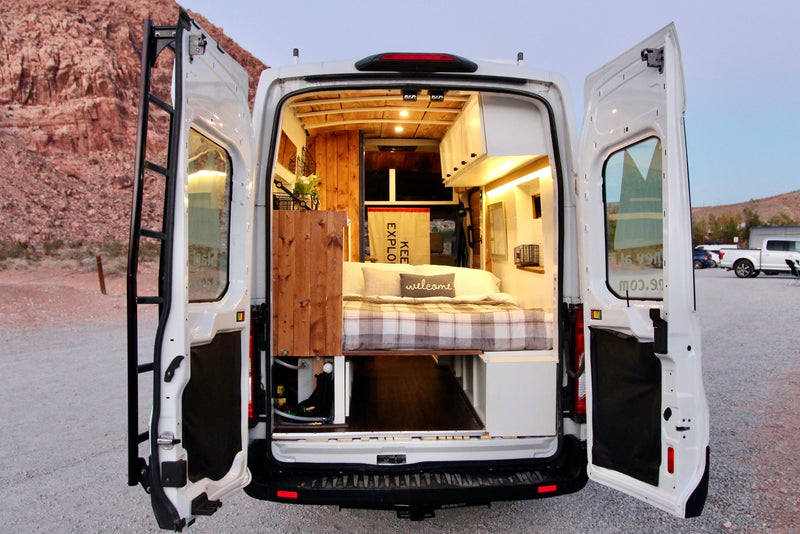 Open rear doors of an RV rental van reveal a cozy interior with a plaid bed, wood paneling, and storage. Parked in a desert setting. — photo 2