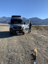 RV rental trip: Silver camper van, person photographing desert mountains, and dog in foreground on dirt road.