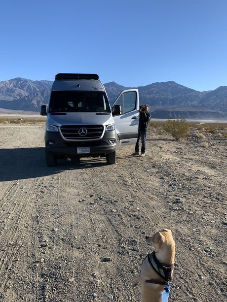 RV rental trip: Silver camper van, person photographing desert mountains, and dog in foreground on dirt road.