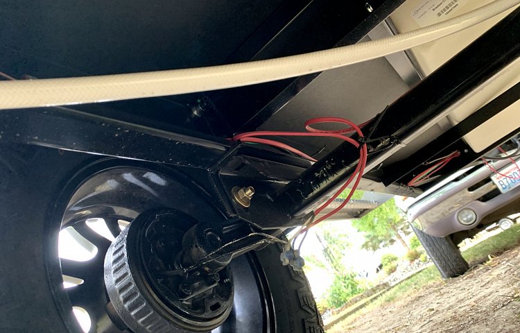 Underside view of an RV rental, showing the black metal chassis, axle, wheel, a white hose, and red electrical wiring. — photo 18