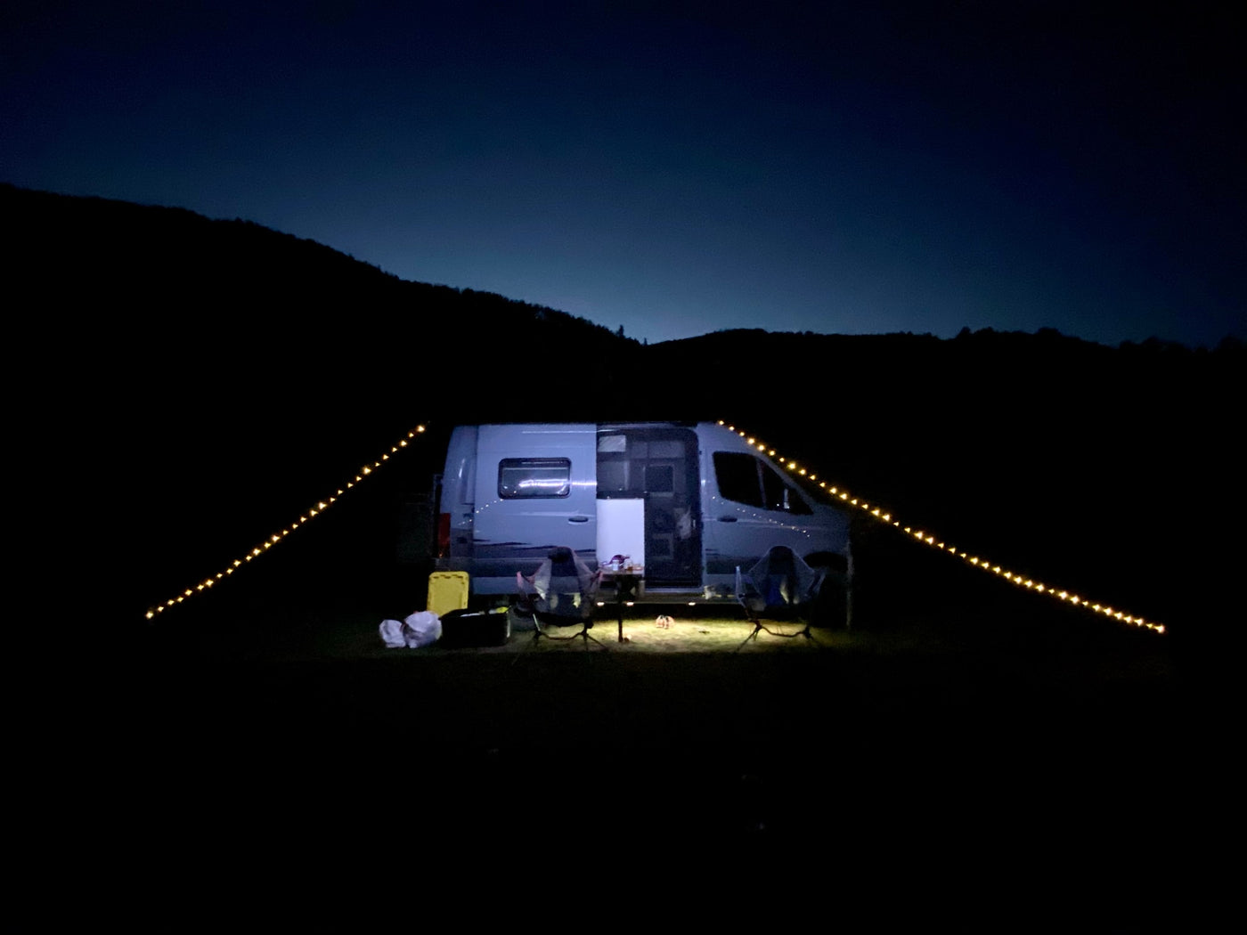 A white camper van at night, illuminated by string lights, with chairs set outside for a cozy RV rental camping experience. — photo 10