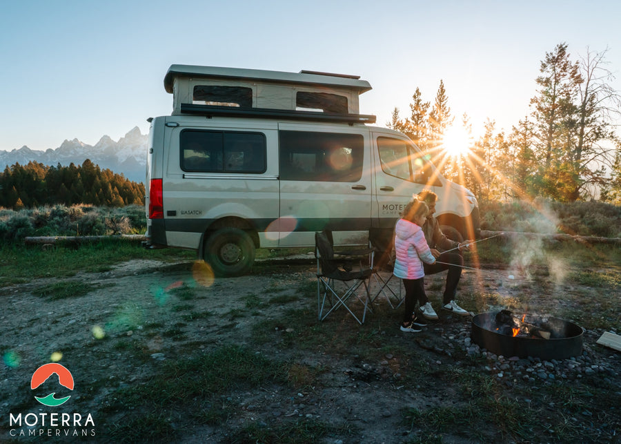 RV rental adventure: Family roasting marshmallows by campfire near their camper van with mountains at sunset.