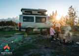 RV rental adventure: Family roasting marshmallows by campfire near their camper van with mountains at sunset.
