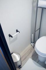 Inside an RV rental bathroom: toilet, toilet brush, empty paper holder, trash can, and towel rack on wood-look floor.