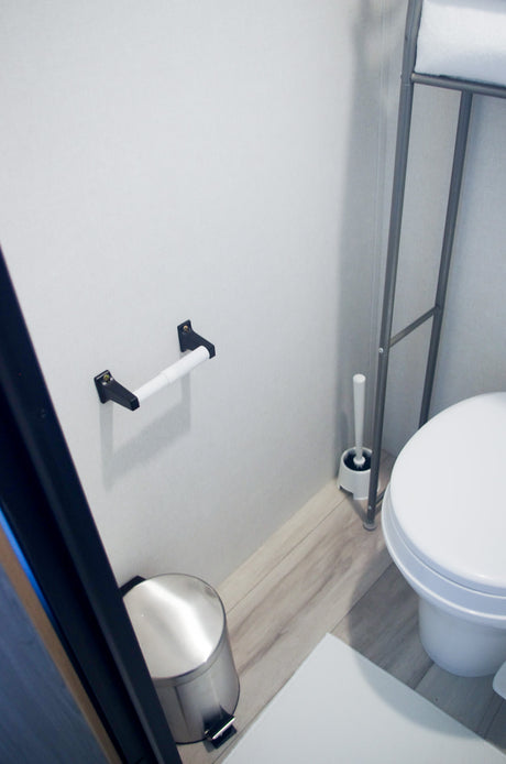 Inside an RV rental bathroom: toilet, toilet brush, empty paper holder, trash can, and towel rack on wood-look floor.