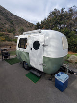 Explore nature with this vintage-style white and green RV rental parked at a serene campsite with a steep hillside and trees.