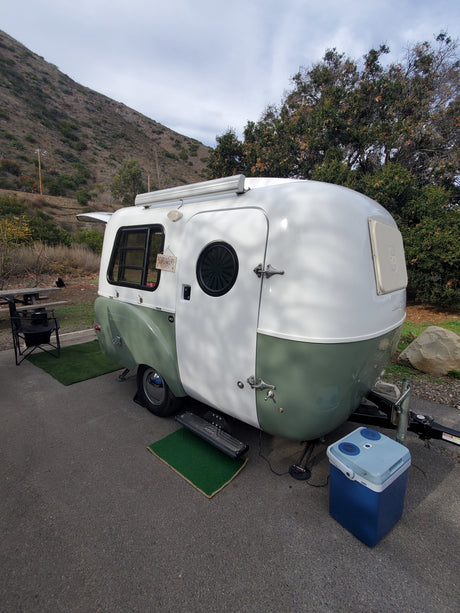 Explore nature with this vintage-style white and green RV rental parked at a serene campsite with a steep hillside and trees.