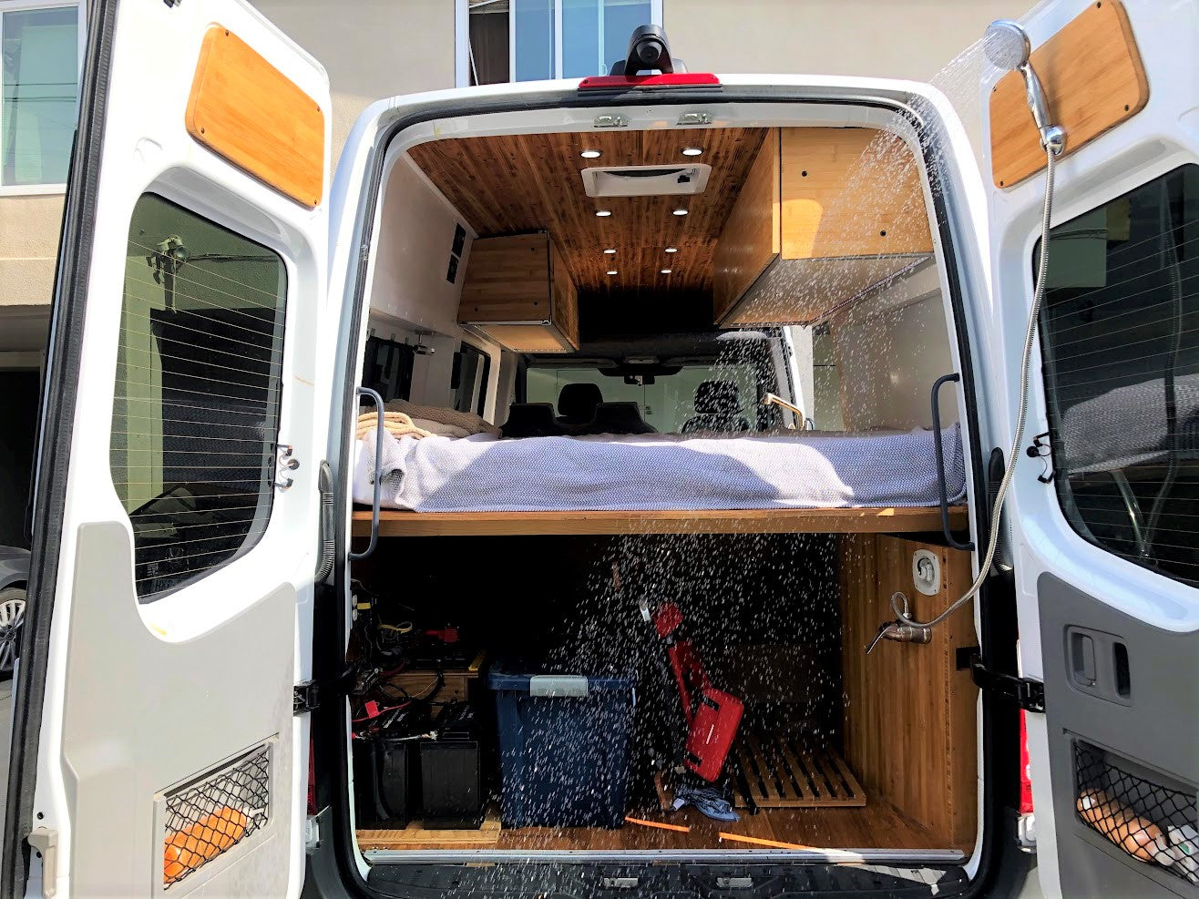 Back of an rv rental camper van. Features a cozy bed, ample storage, and an outdoor shower spraying water.