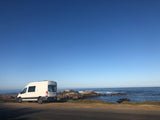 White RV rental van parked by a rocky ocean coast under a clear blue sky with a faint moon. Road trip adventures await!