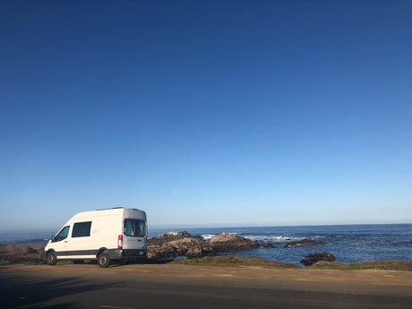 White RV rental van parked by a rocky ocean coast under a clear blue sky with a faint moon. Road trip adventures await!