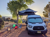 RV rental van with rooftop tent set up at a peaceful campsite, surrounded by chairs & a tree, under a serene sunset.