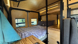 Modern RV rental interior with a cozy bed, wood paneling, and thoughtful storage, bathed in ambient light.