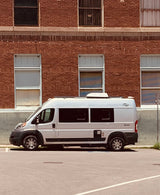 A white rv rental camper van is parked by a brick building, its compact size ready for travel adventures.