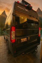 Rear view of a dark Jayco RV rental camper van at sunset, parked lakeside with mountains in the background.