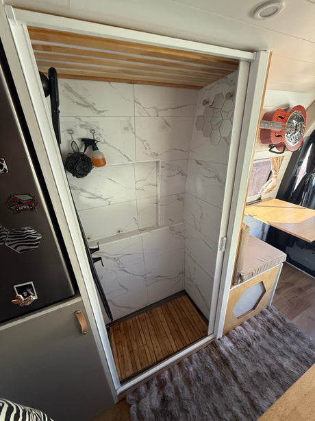 This rv rental features a compact shower with elegant marble-look tiles, a wooden slatted floor, and storage for toiletries.