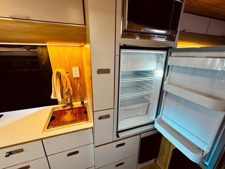 Compact RV rental interior featuring a functional kitchen with sink, microwave, and an empty, open refrigerator.
