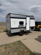 Compact Coleman travel trailer RV rental with a yellow Jeep, parked on concrete under a cloudy sky.