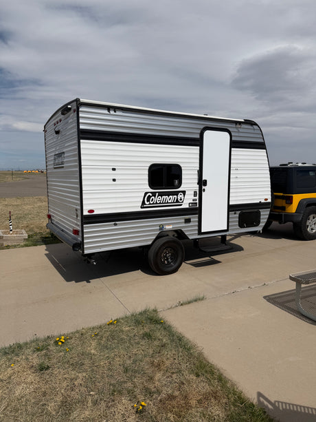 Compact Coleman travel trailer RV rental with a yellow Jeep, parked on concrete under a cloudy sky.