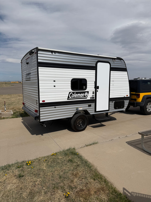 Compact Coleman travel trailer RV rental with a yellow Jeep, parked on concrete under a cloudy sky.