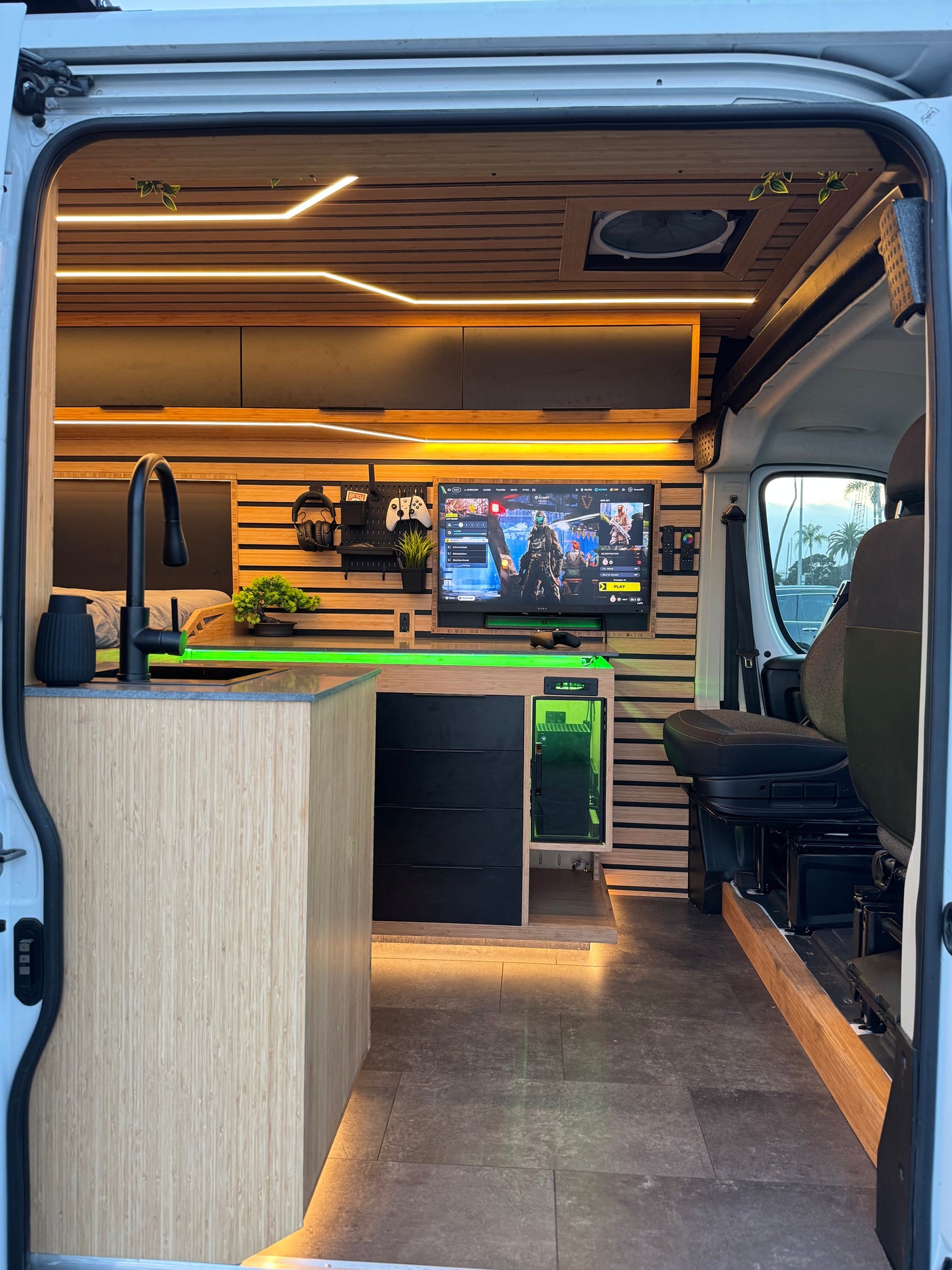 Stylish RV rental interior: gaming setup, kitchen, bed, slatted wood walls, and vibrant green LEDs for a modern mobile retreat.