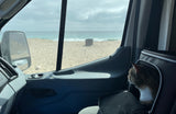 Cat in RV rental carrier gazes at ocean waves and sandy beach from inside the vehicle, with a clear horizon.
