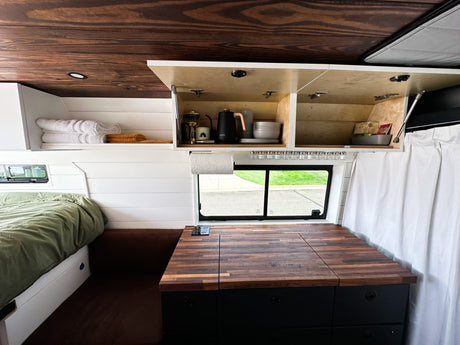 This RV rental showcases a cozy van interior with a bed, shiplap walls, wood ceiling, and a functional kitchen with opened cabinets.