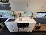 RV rental interior featuring a compact kitchen with sink, microwave, and storage, next to a comfy bed with windows.